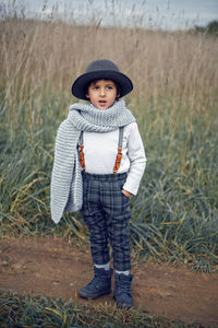 Boy child in plaid pants, hat, suspenders and scarf stands in a field in autumn