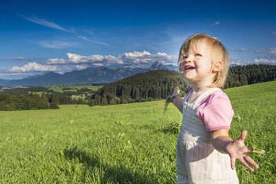 Smiling cute girl gesturing while standing on grass against sky
