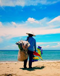 Rear view of senior man selling kites while standing at beach against sky during sunny day