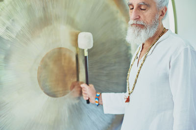 Side view of crop adult male in white clothes with mallet playing suspended gong during spiritual practice