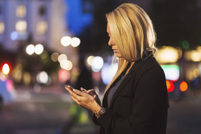 Side view of young woman using smart phone on city street at night