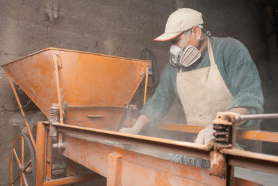 Male worker in apron and protective respirator cleaning plaster station with brushes while working in professional ceramic workshop