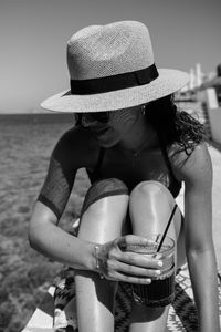 Young woman wearing hat sitting at beach with drink during sunny day