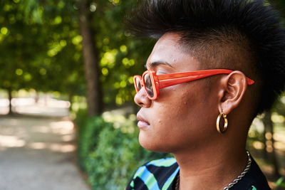 Filipino non binary person in stylish sunglasses with acne on face looking away while standing near tropical lush bush on summer day in park