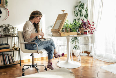 Artist mixing colors in palette sitting on chair in front of easel at home