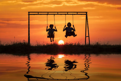 Silhouette siblings swinging over lake against sky during sunset