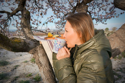 Young blond woman having hot tea almond blooming trees early morning 