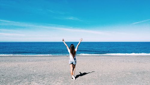 Rear view of woman with arms raised enjoying at beach against sky on sunny day