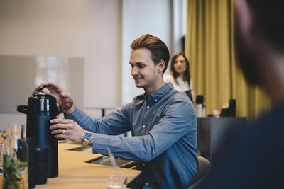 Smiling businessman opening insulated drink container at conference table with colleagues in board room