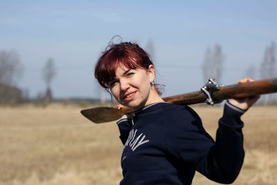 Portrait of young woman holding oar while standing on field against clear sky