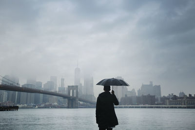 Rear view of man holding umbrella while looking at city view against cloudy sky