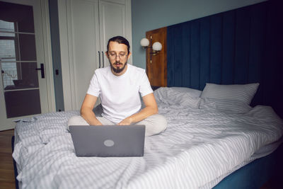 Man in a white t-shirt, a freelancer with glasses, sits on the bed at home with a laptop and works 