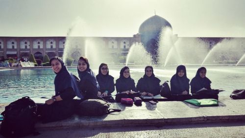 People sitting on fountain in city against sky