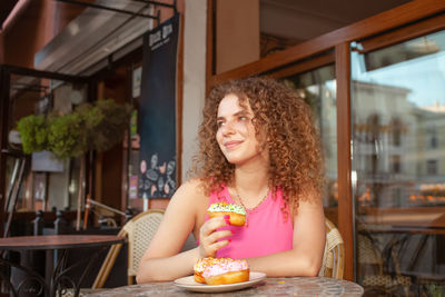Portrait of beautiful curly young woman in cafe on summer terrace, delicious colorful donuts,
