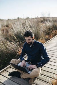 Man using laptop while sitting on boardwalk against sky