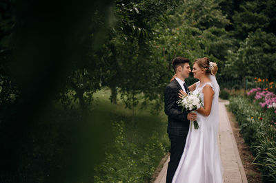 Newlywed couple looking at each other while standing against trees