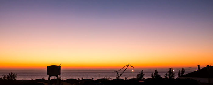 Silhouette storage tank and crane by sea against clear sky during sunset