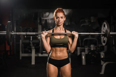 Mid adult woman looking at camera holding barbell olympic bar in gym during intense workout