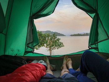 Low section of people relaxing by lake against sky