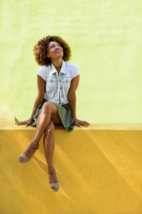 Young woman looking up while sitting against yellow wall