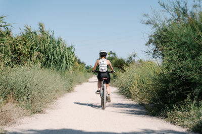 Full body of positive young female cyclist in sportswear and helmet riding bicycle on dirt path among green plants in summer day in countryside