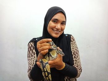 Portrait of smiling young woman holding snake against white wall