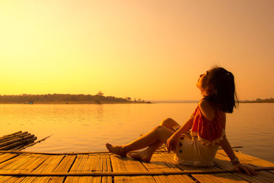 Woman sitting on wooden raft in lake against clear sky during sunset