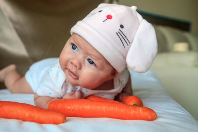 Close-up of cute baby girl wearing bunny hat while lying with carrots on bed