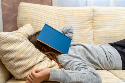 Girl with book on face relaxing over sofa at home