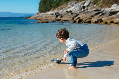 Side view of little preschool boy in denim overall standing barefoot on wet sand and sailing toy ship on clear blue water