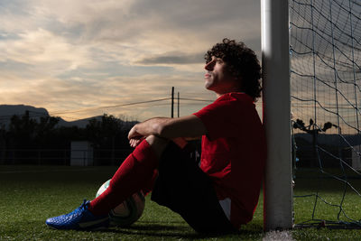 Full body side view of pensive soccer player with ball sitting near goal on sports field and looking away in evening