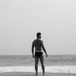 Rear view of shirtless muscular man standing on shore at beach against clear sky