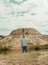 A man standing infront of water looking at a big rock object