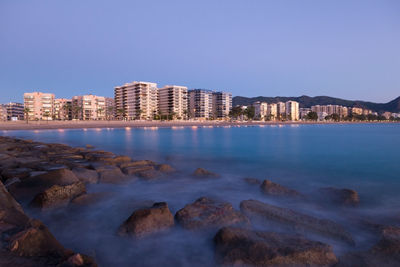 Sea by illuminated city against clear blue sky