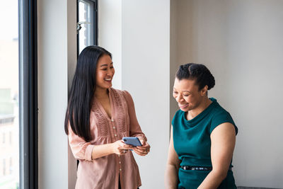 Smiling businesswoman showing mobile phone to female colleague by window at office