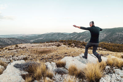 Back view full body of unrecognizable male spreading arms while jumping on stones in hilly rocky terrain during adventure