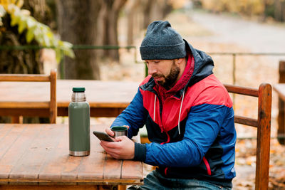 A young man using a mobile phone is sitting outdoors in a cafe or restaurant on an autumn day