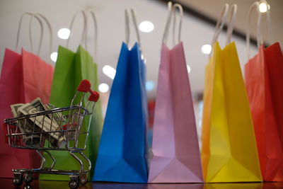 Close-up of colorful shopping bags with paper currencies in miniature shopping cart on wooden table