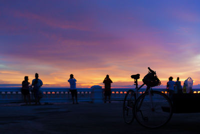Silhouette people riding bicycles on sea against sky during sunset