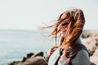 Side view of happy woman with tousled hair standing on pier over sea against clear sky