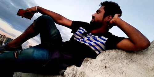 Young man sitting on rock against sky