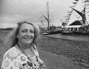 Portrait of smiling mature woman standing on field against moored boats at harbor