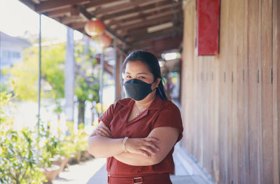 An asian woman in a brown shirt wearing a face mask crossed her arms looking at a camera 