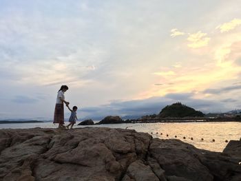 Mother and daughter holding hands while standing on rocks against sky during sunset