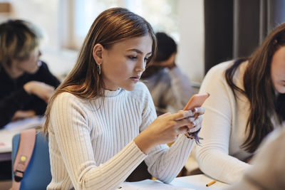 Teenage girl with brown hair using smart phone sitting by female friend at desk in classroom