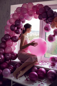 Brunette woman in a purple dress sits on a window with pink balloons at a birthday party