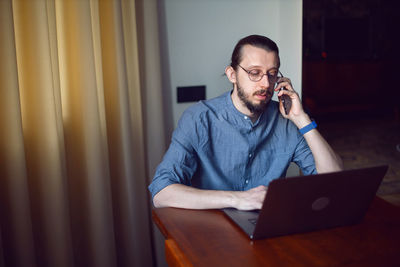 Businessman with a beard and glasses is sitting at a laptop at a wooden table. talking on the phone