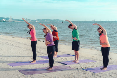 Young friends stretching arms while standing on shore at beach during sunny day