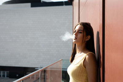 Young woman smoking while standing by wall