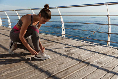 Full length of woman tying shoelace on floor against sea and sky